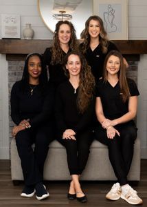Five women seated and standing in front of a fireplace, dressed in black and smiling.