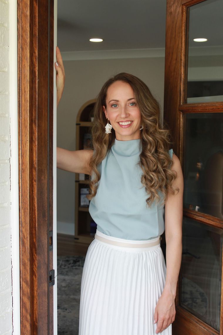 Woman standing in a doorway, smiling, wearing a light blue top and white pleated skirt.