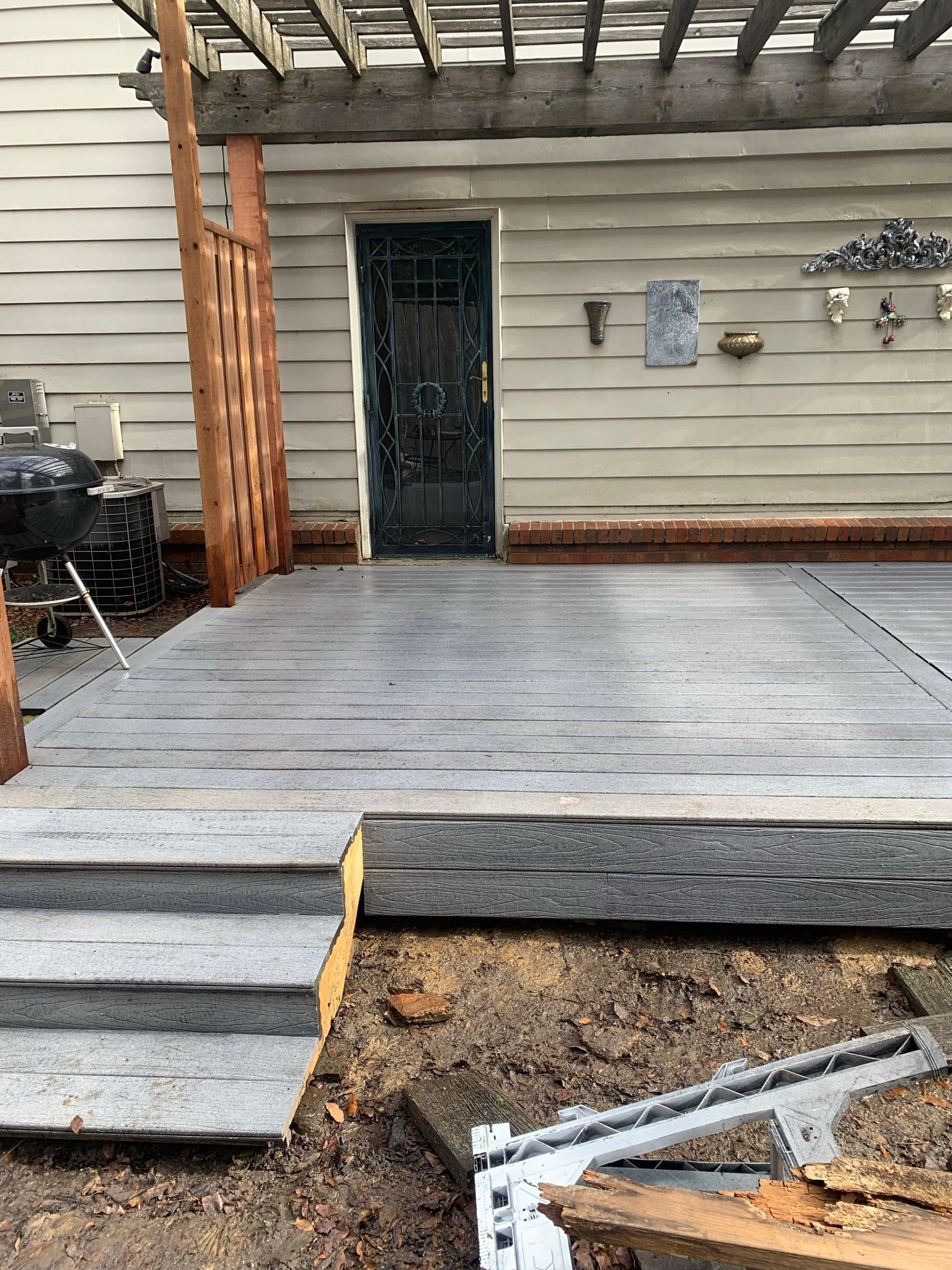 Grey concrete deck with steps leading to a door, pergola overhead. Brown wood beams and siding.