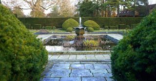 There is a fountain in the middle of the garden surrounded by bushes.