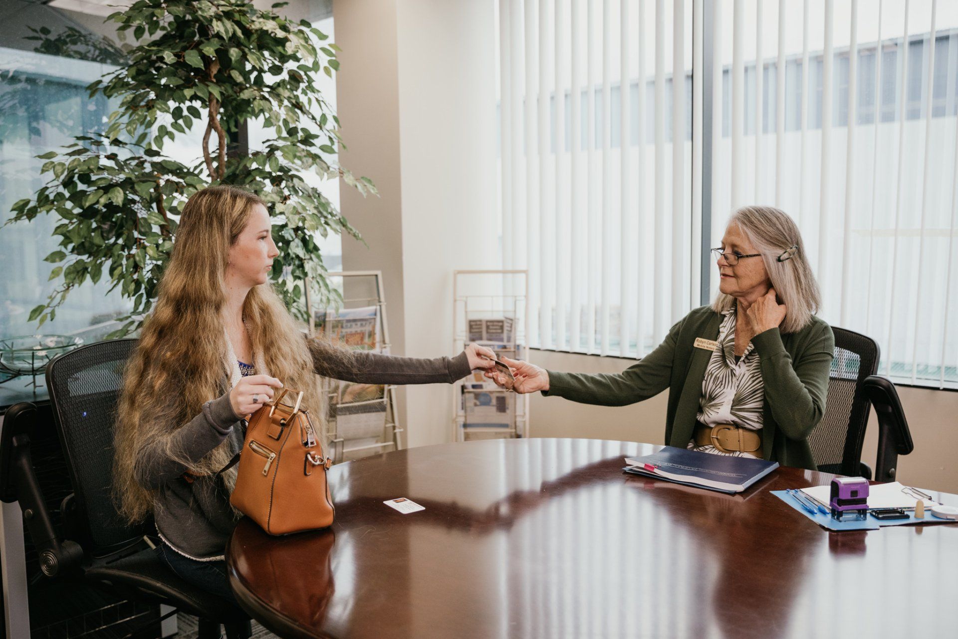 Two women seated at a conference table exchange a document in a bright office.