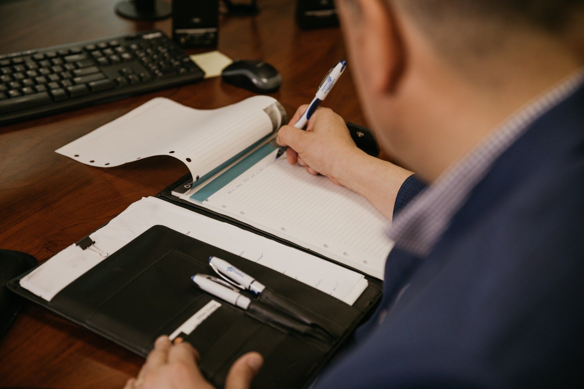 Man writing in notebook at desk, open binder with two pens, keyboard and mouse visible.
