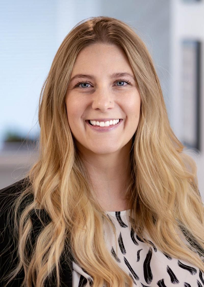 Woman with long blonde hair smiles, wearing a patterned top and black blazer in an office setting.