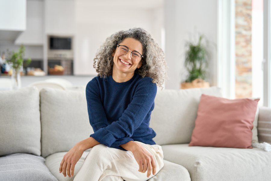 Woman with curly gray hair, smiling on a sofa in a living room.