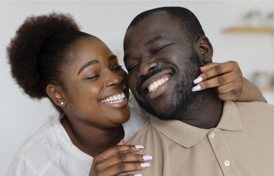 Smiling couple embracing, indoors; woman's arm around man's neck. They both have closed eyes, and are laughing.