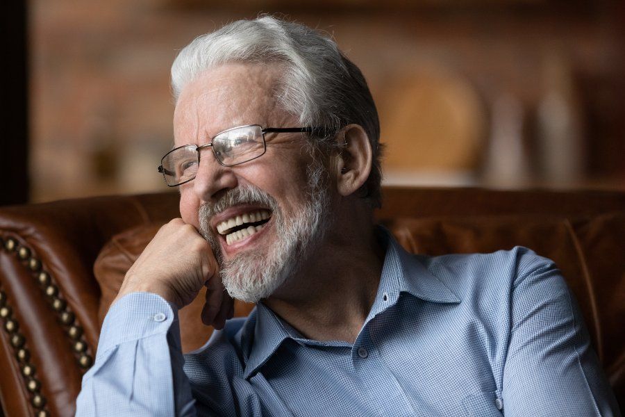 Smiling person with gray hair and beard, wearing glasses, in a blue shirt, sitting in a leather chair.