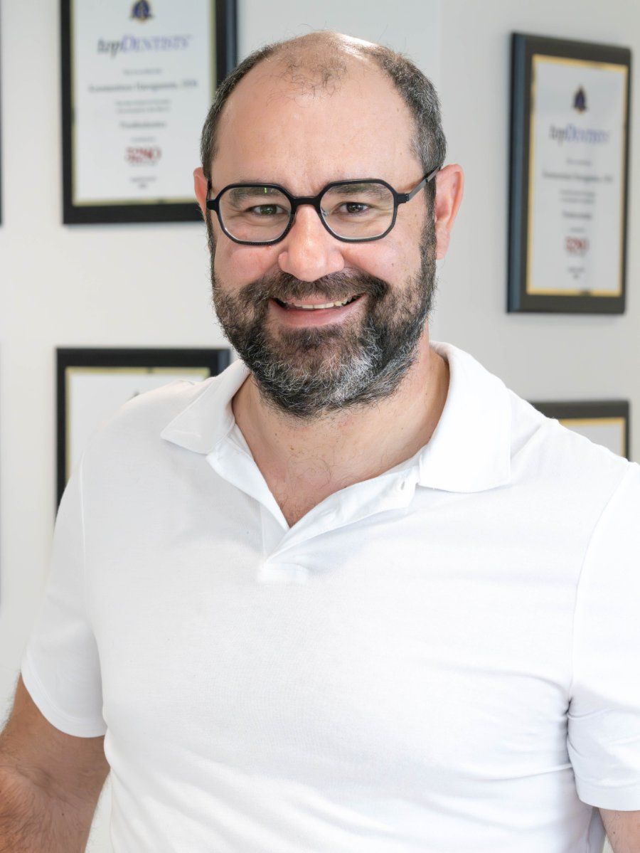 Man wearing glasses, white shirt, smiling, in front of framed documents.
