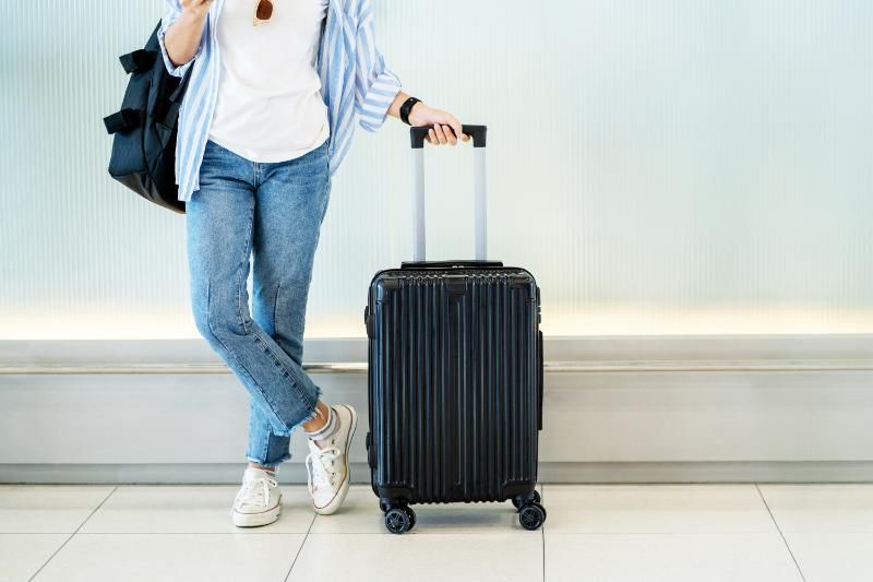 Woman leaning on a black rolling suitcase, wearing jeans, shirt, and backpack in a brightly lit indoor space.