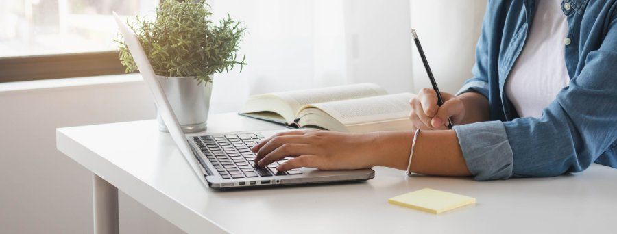 Person writing on a laptop with a pencil and open book on a white desk by a window.