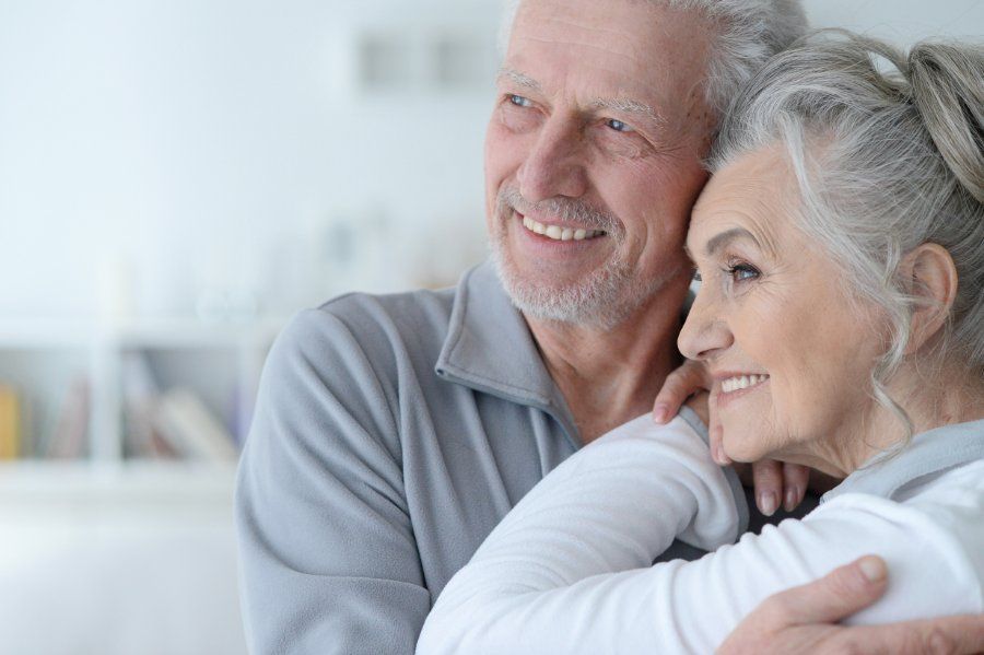 Older couple embracing, smiling, looking away, in a brightly lit room.