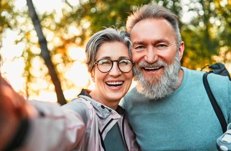 Smiling couple taking a selfie outdoors; woman in glasses, man with beard, trees in background.