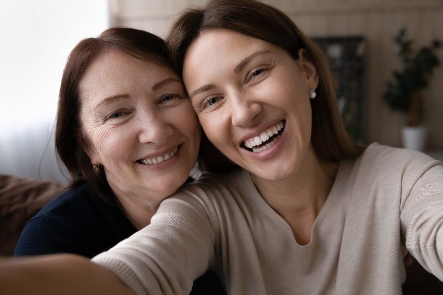 Woman and older woman smiling, taking a selfie. Indoors, casual setting.