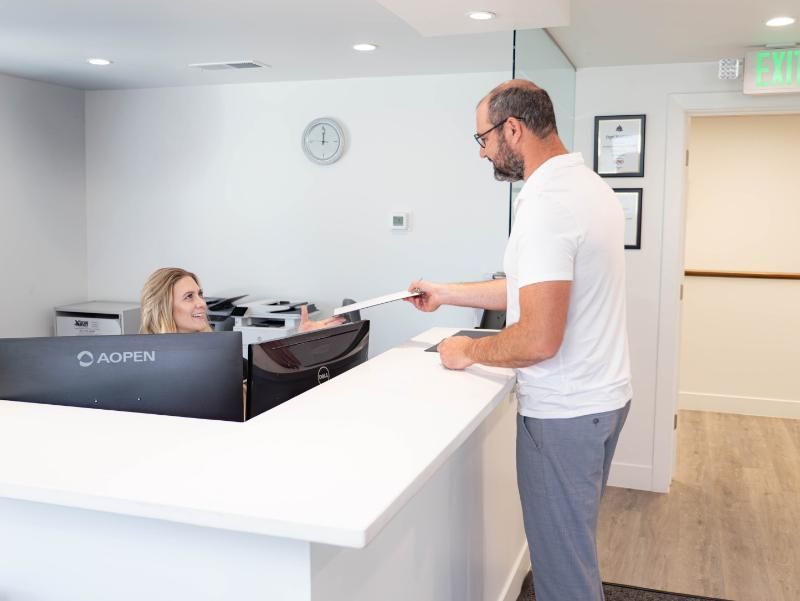 Man handing papers to receptionist at white front desk in a medical office.