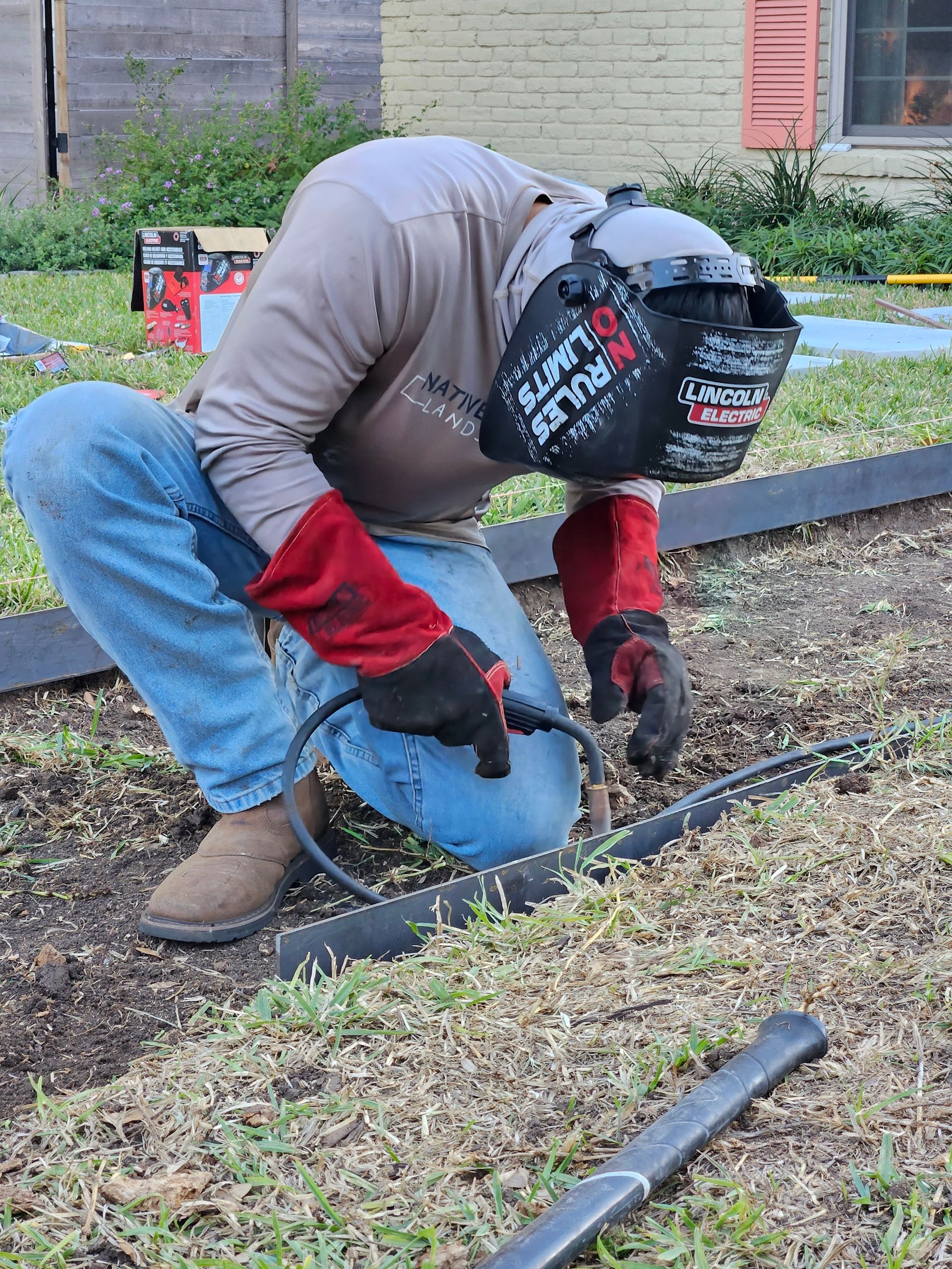 A man wearing a welding helmet is kneeling down in the grass.