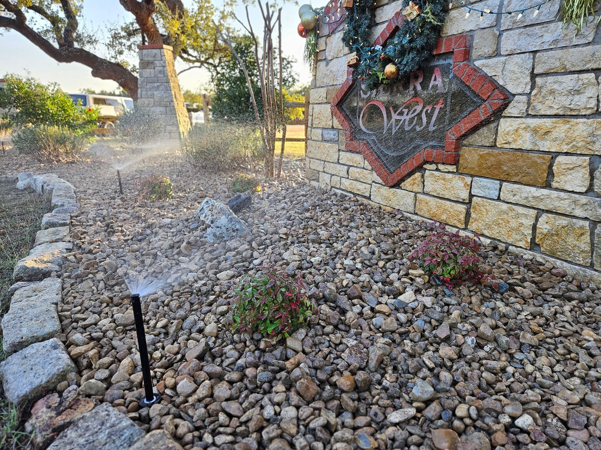 A sprinkler is spraying water on a rocky area in front of a brick wall.