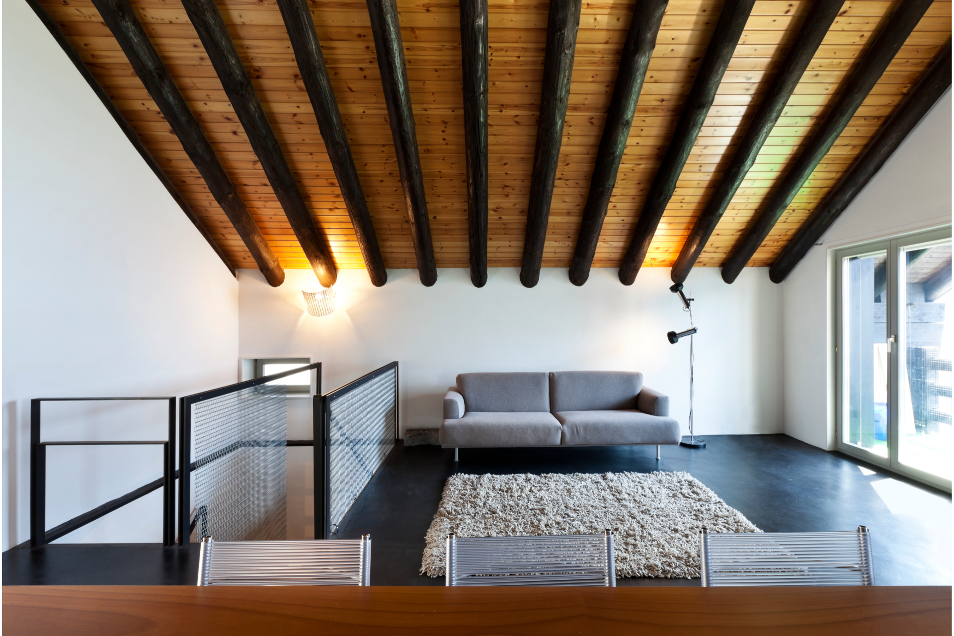 Loft living room with wood beam ceiling, gray sofa, white walls, and dark floor.