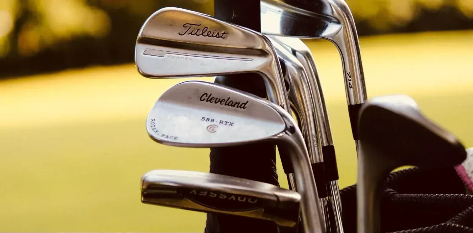 A set of golf clubs, including wedges and a putter, standing in a bag on a blurred golf course background.