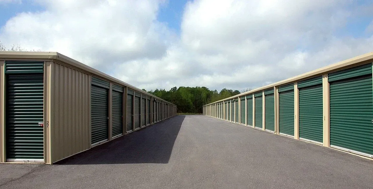 Two rows of beige self-storage units with dark green roll-up doors line a gravel path under a cloudy sky.