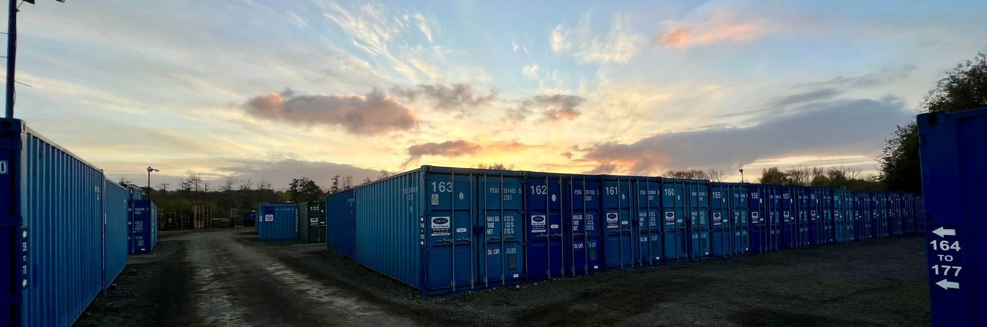 A row of blue shipping containers stands in an outdoor storage yard at sunset with a golden, cloudy sky in the background.
