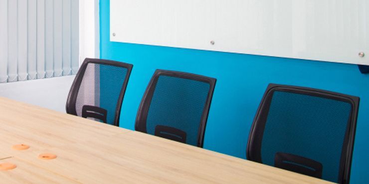 Three black office chairs sit along a light wood table against a bright blue wall with a whiteboard above.