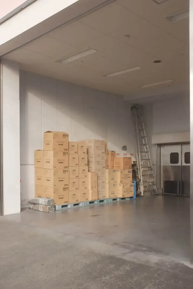 A stack of cardboard boxes on a blue pallet against a white wall in a large, open industrial room with a tall ladder.