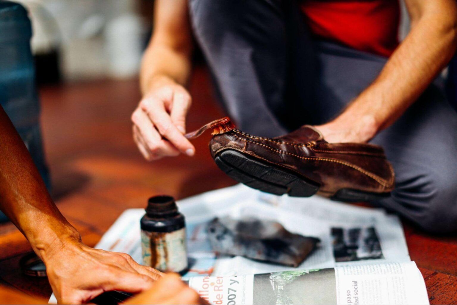 A person uses a small brush to apply brown polish to a dark brown leather loafer resting on a newspaper-covered floor.