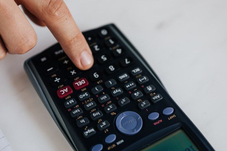 A person’s finger presses a button on a black scientific calculator resting on a white surface.