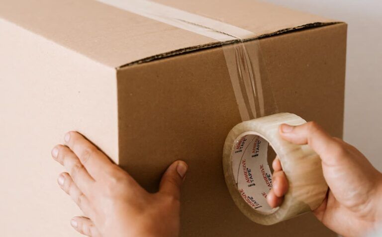 Hands securing a cardboard box with a roll of clear packing tape.