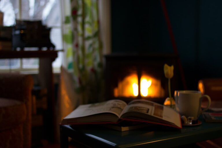 An open book and a white mug sit on a table in front of a warm fireplace in a cozy, dimly lit room.