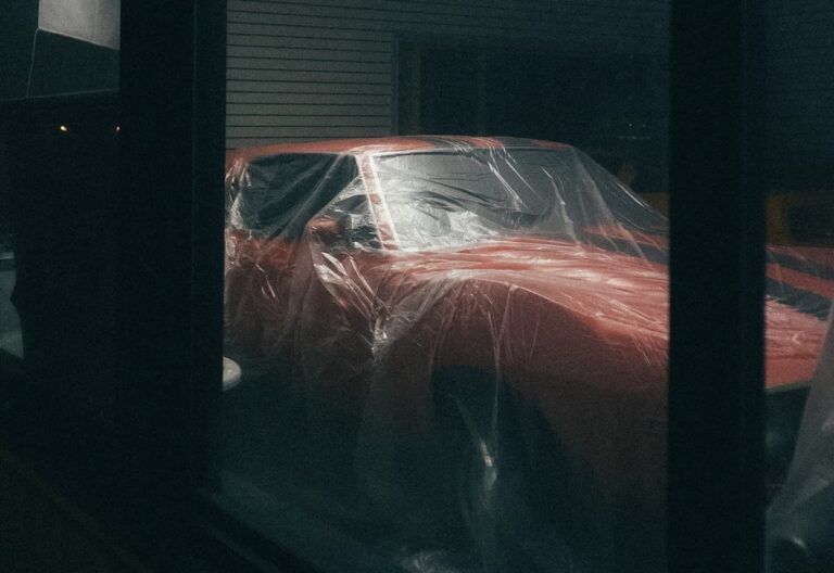 A red car with black stripes is parked indoors and draped in clear plastic sheeting, viewed through a window.