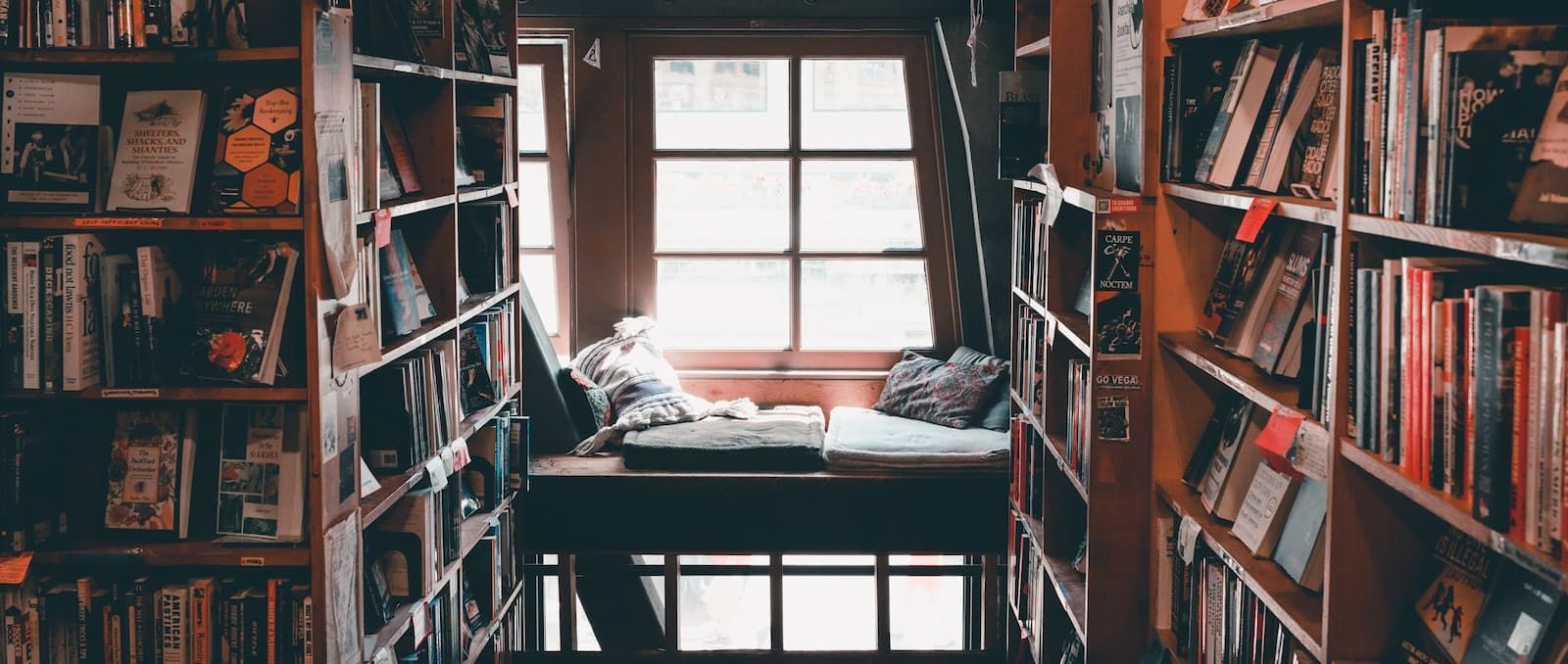 A cozy bookstore nook with a window seat between tall wooden shelves packed with books.