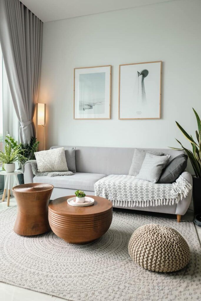 A modern living room with a grey sofa, two wooden coffee tables, a woven pouf, and two framed prints on a white wall.