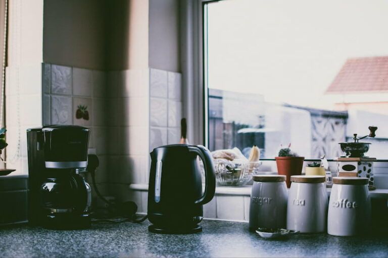A coffee maker, black electric kettle, and three labeled storage jars sit on a kitchen counter in front of a window.