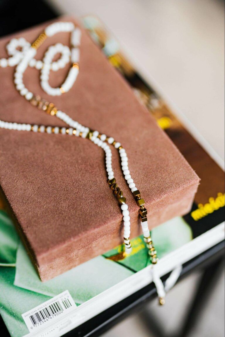 A beaded necklace with white pearls and gold accents rests on top of a soft brown box sitting on a magazine.
