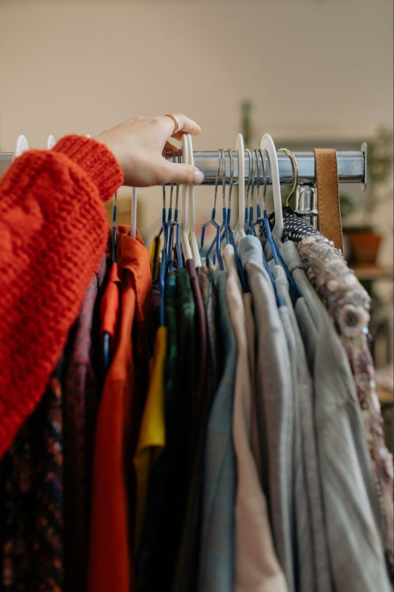 A hand wearing a bright red sweater selects a garment from a clothing rack filled with various items.