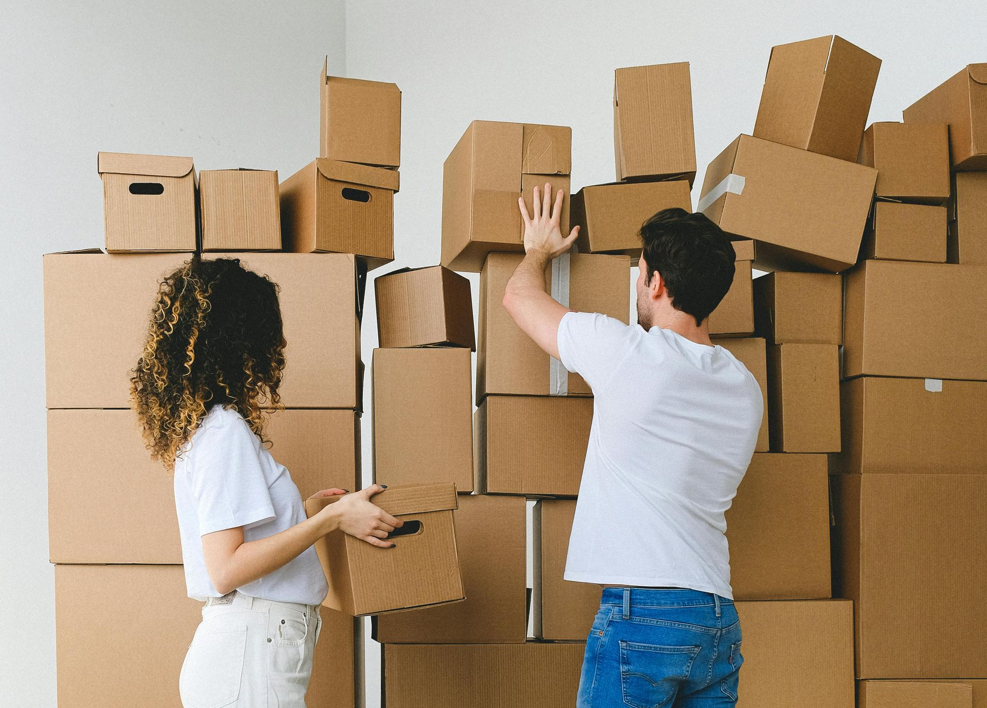 A man and woman are stacking various cardboard moving boxes against a white wall in a bright room.