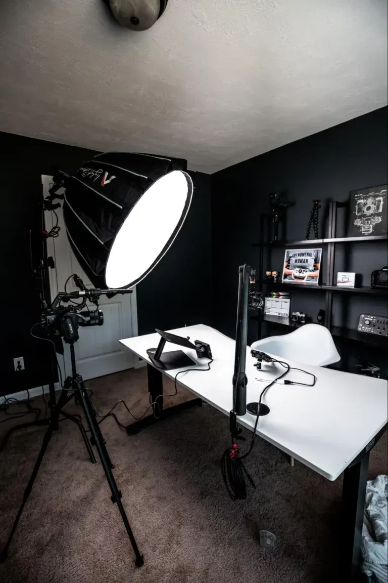 A bright studio light on a tripod illuminates a white desk with tech gear against a dark wall in a home office.