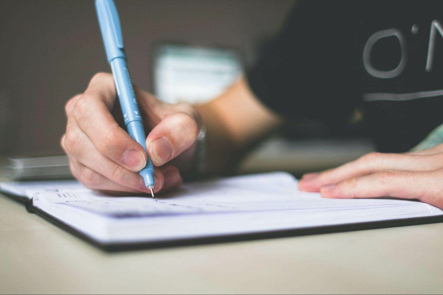 A hand writes in an open notebook with a light blue pen at a desk.