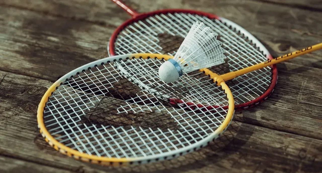 A white shuttlecock resting on two badminton rackets with red and yellow frames, set against a dark wooden surface.