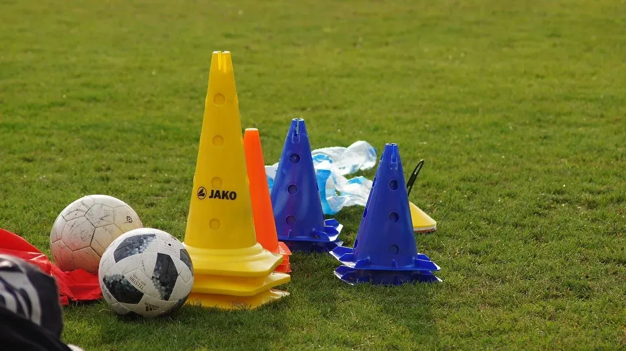 Soccer training equipment, including two soccer balls and several yellow, orange, and blue cones, sits on a grass field.