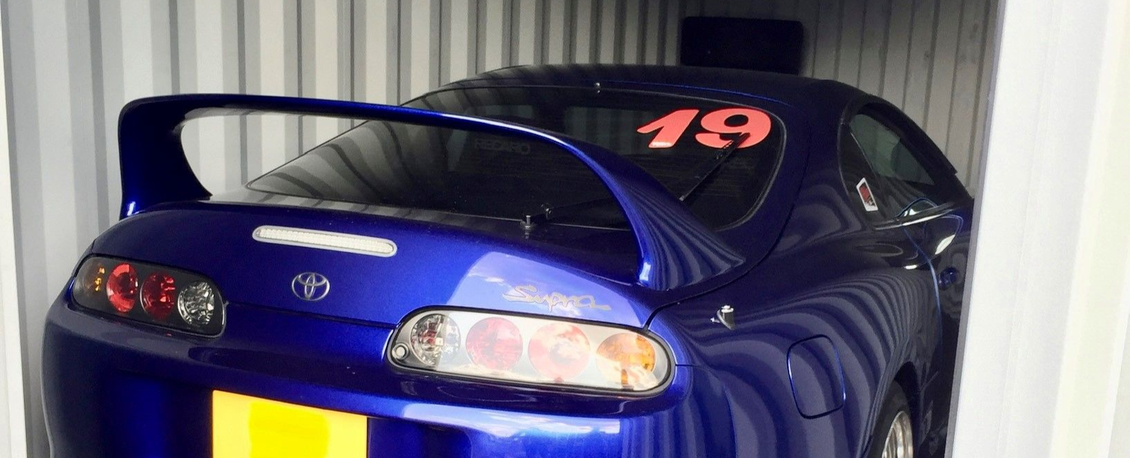 A bright blue Toyota Supra with a racing number 19 on the rear window, parked inside a storage unit.