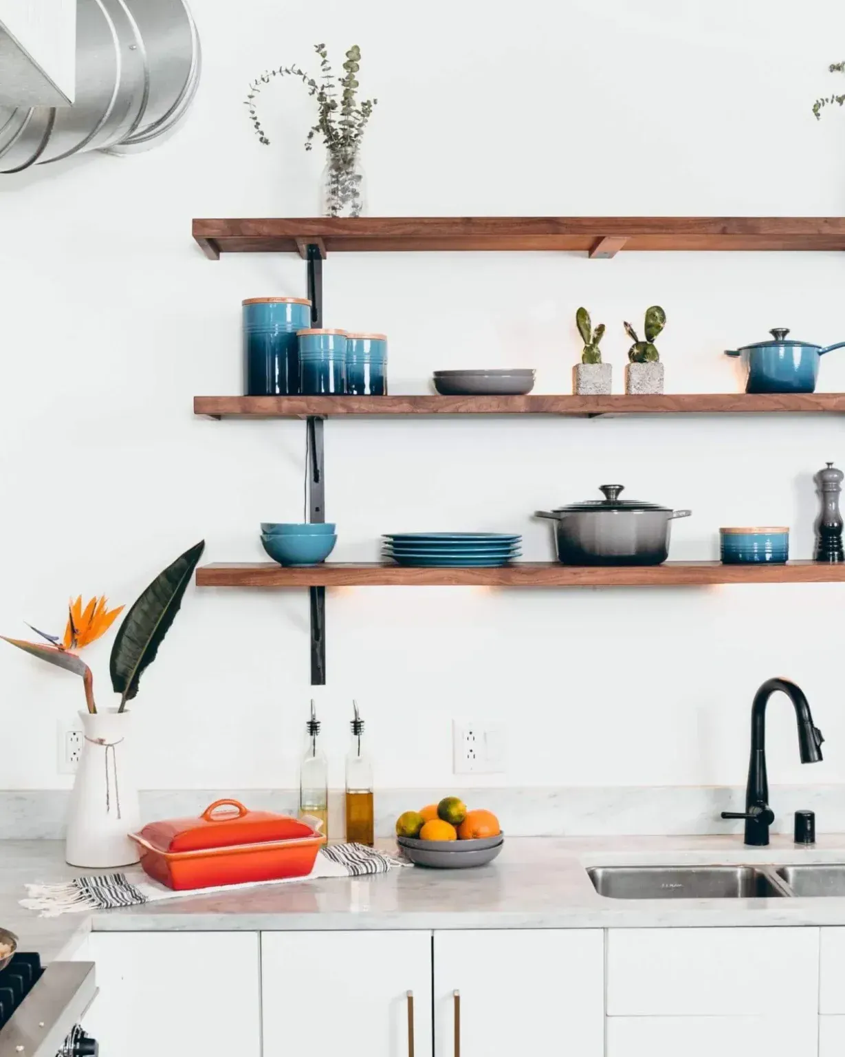 A modern kitchen features white cabinets, wooden open shelving with blue kitchenware, and an orange baking dish on a counter.