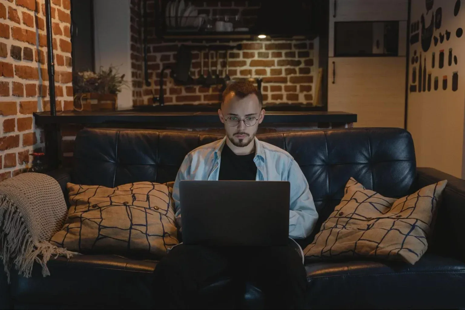 A person in glasses wearing a light-colored shirt sits on a dark sofa, working on a laptop in a brick-walled room.