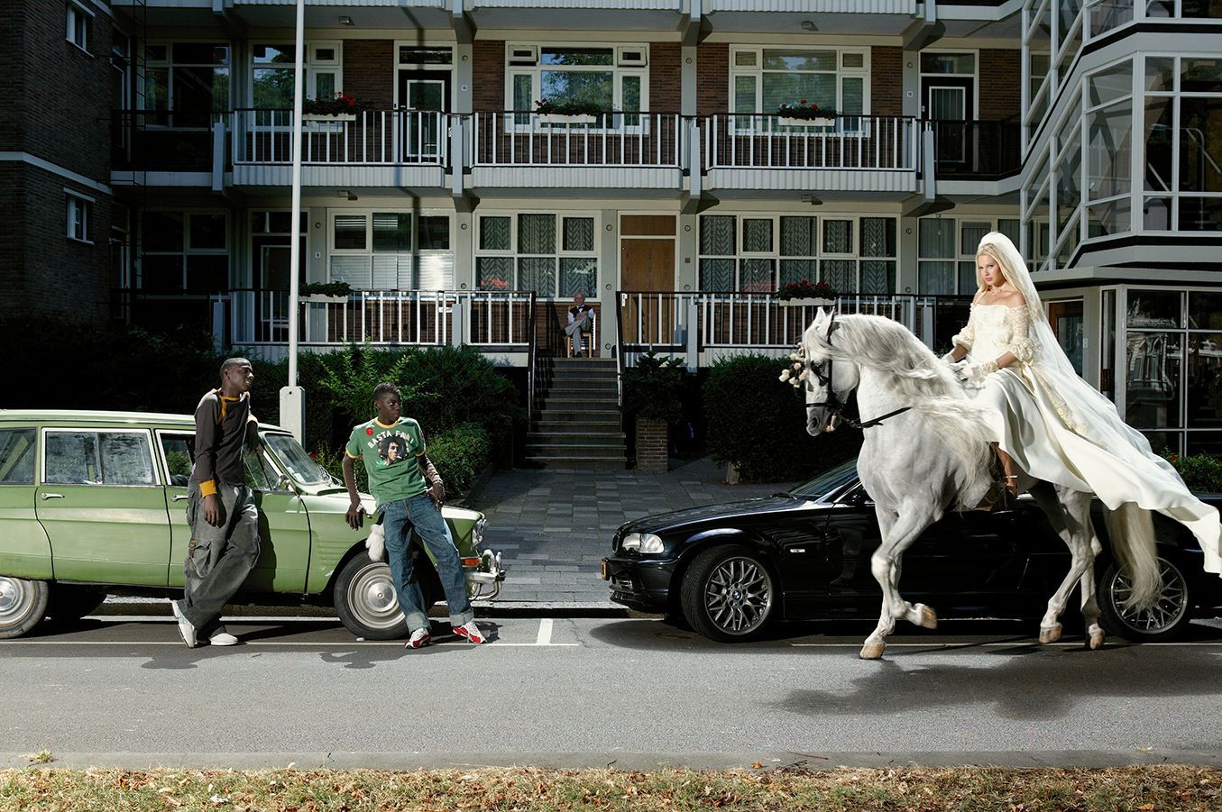 Twee mannen bij een groene auto, een vrouw op een wit paard naast een zwarte auto voor een appartementencomplex.
