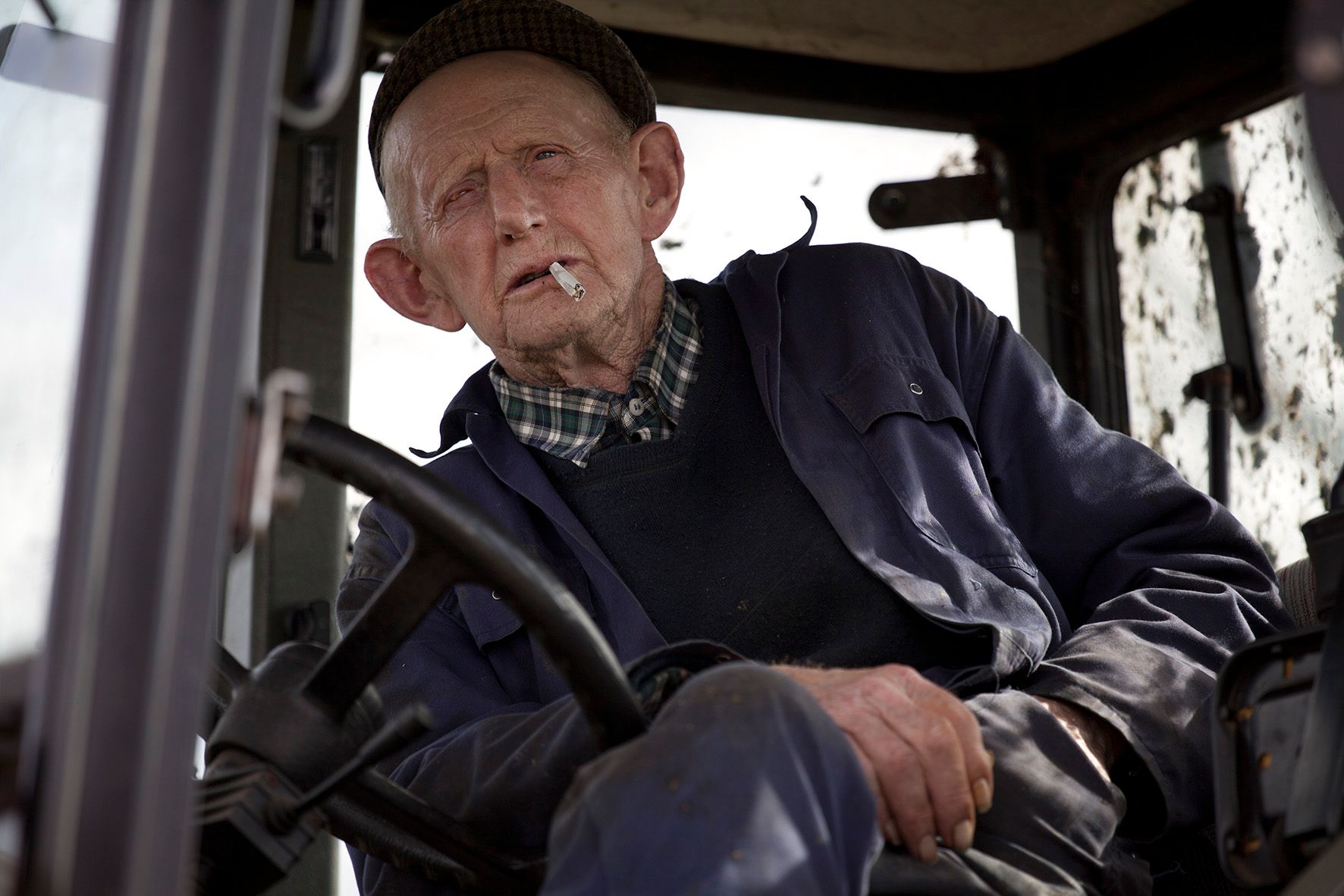 Een oudere boer in een tractor, die een sigaret rookt en vooruitkijkt.