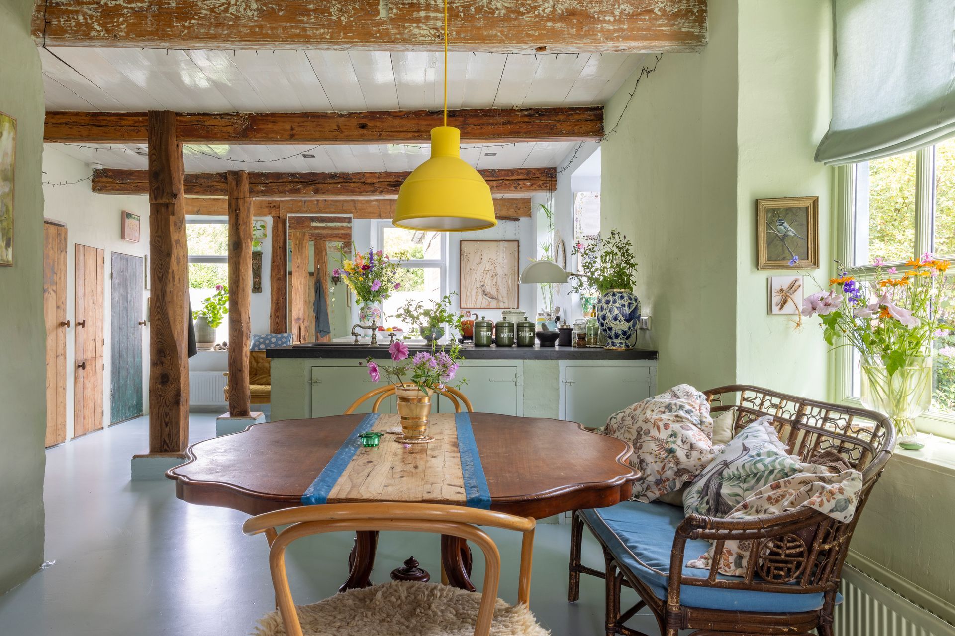 Lichte eetkamer met houten tafel, rieten stoelen, gele hanglamp en bloemen bij de grote ramen, huis van Elise Klinkert