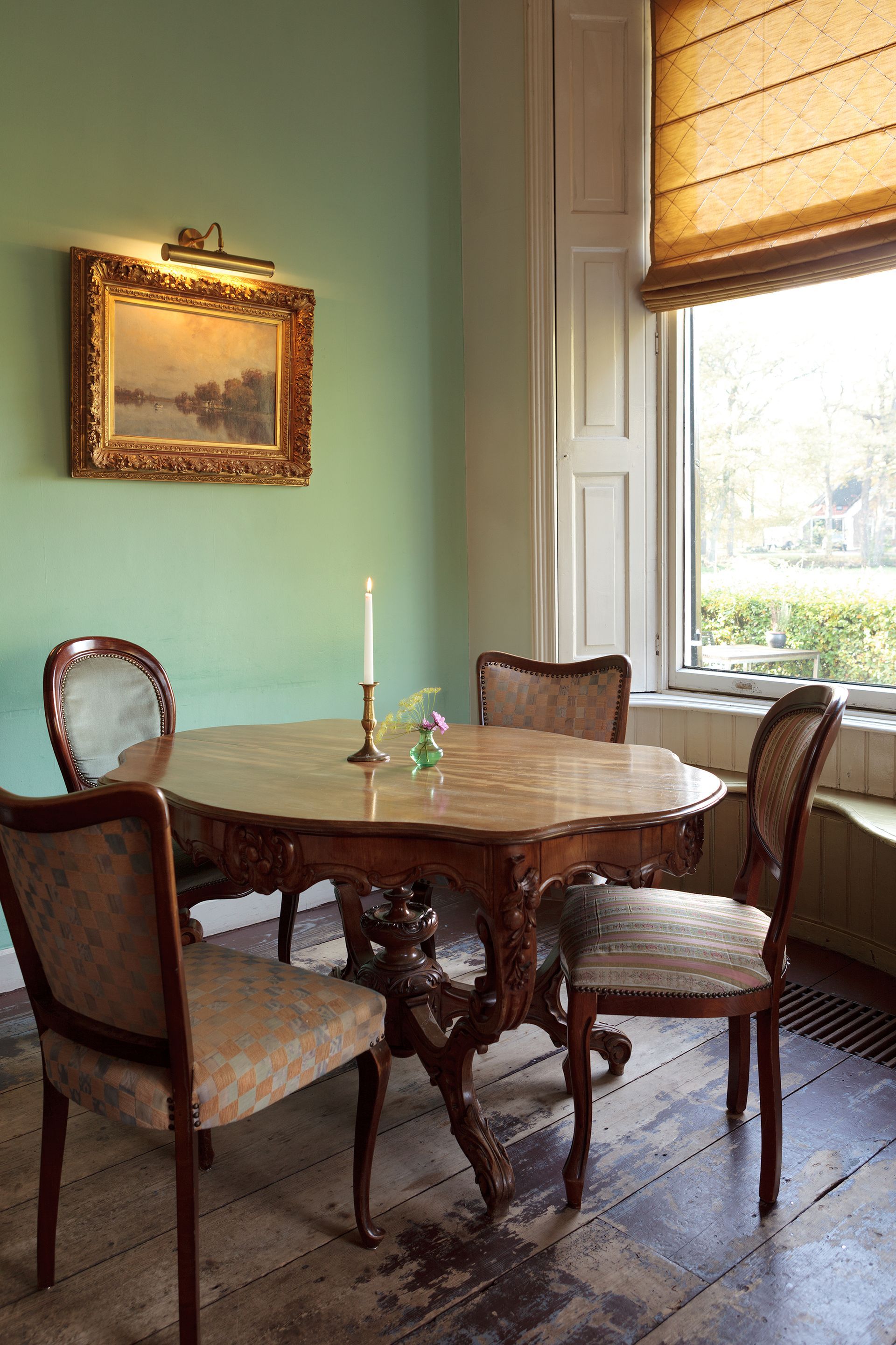 Een eetkamer met een ronde tafel, stoelen, een schilderij en een raam met een zonnescherm.