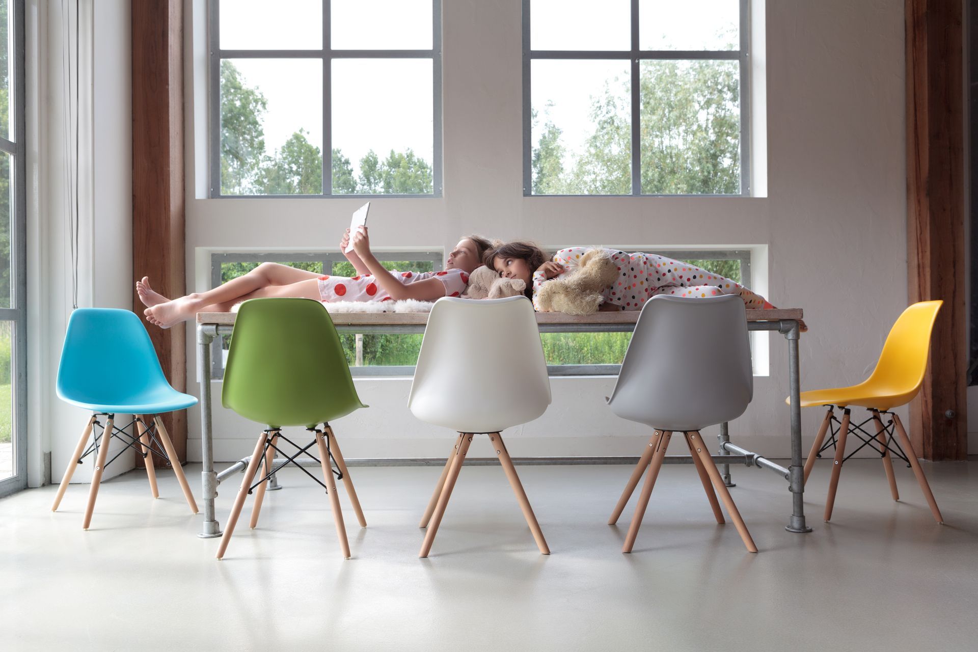 Kinderen liggen op een tafel, omringd door kleurrijke stoelen, in een lichte kamer met grote ramen. Foto Elise Klinkert