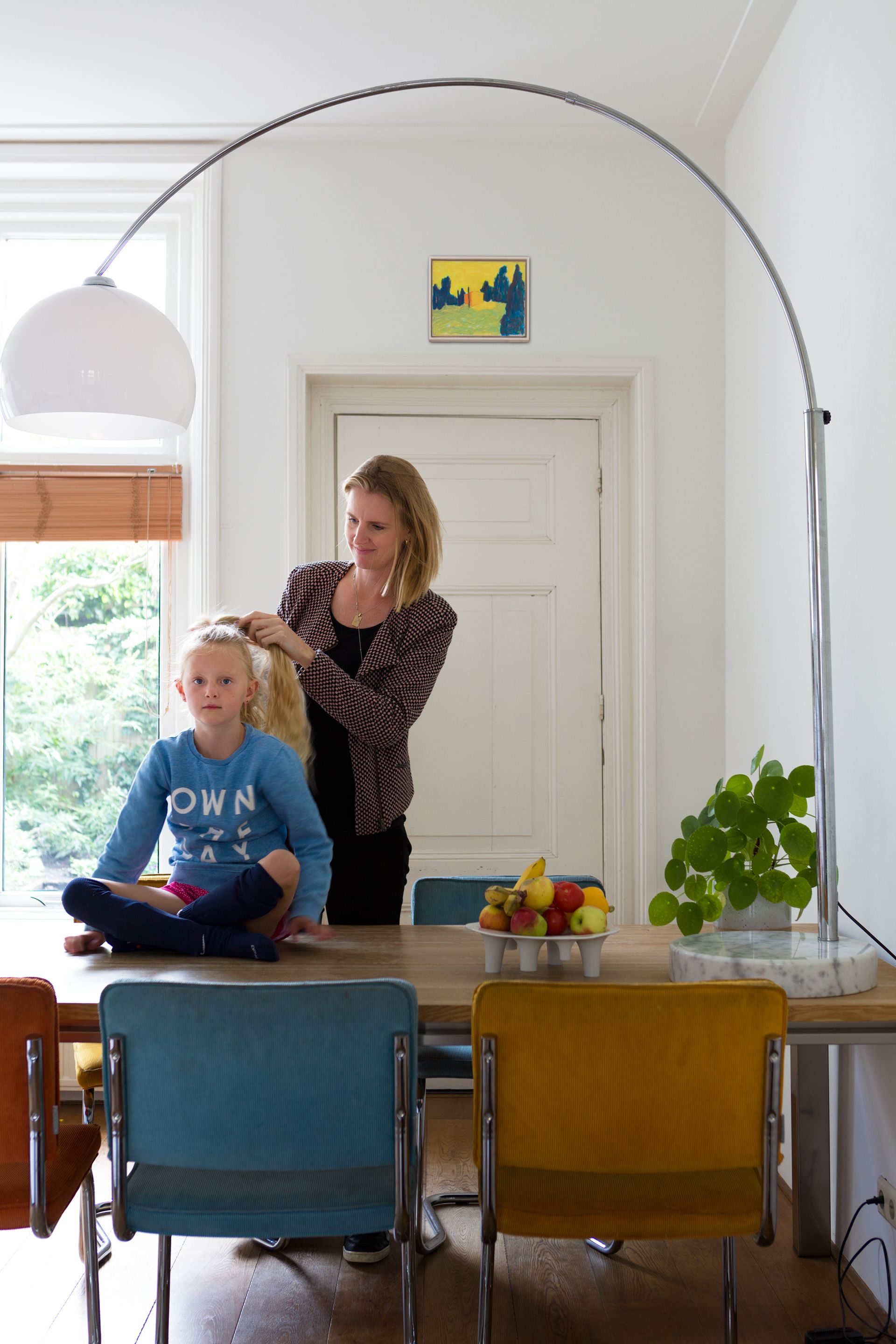 Een vrouw vlecht het haar van een kind aan een tafel. Een grote booglamp hangt boven hen. Foto Elise Klinkert