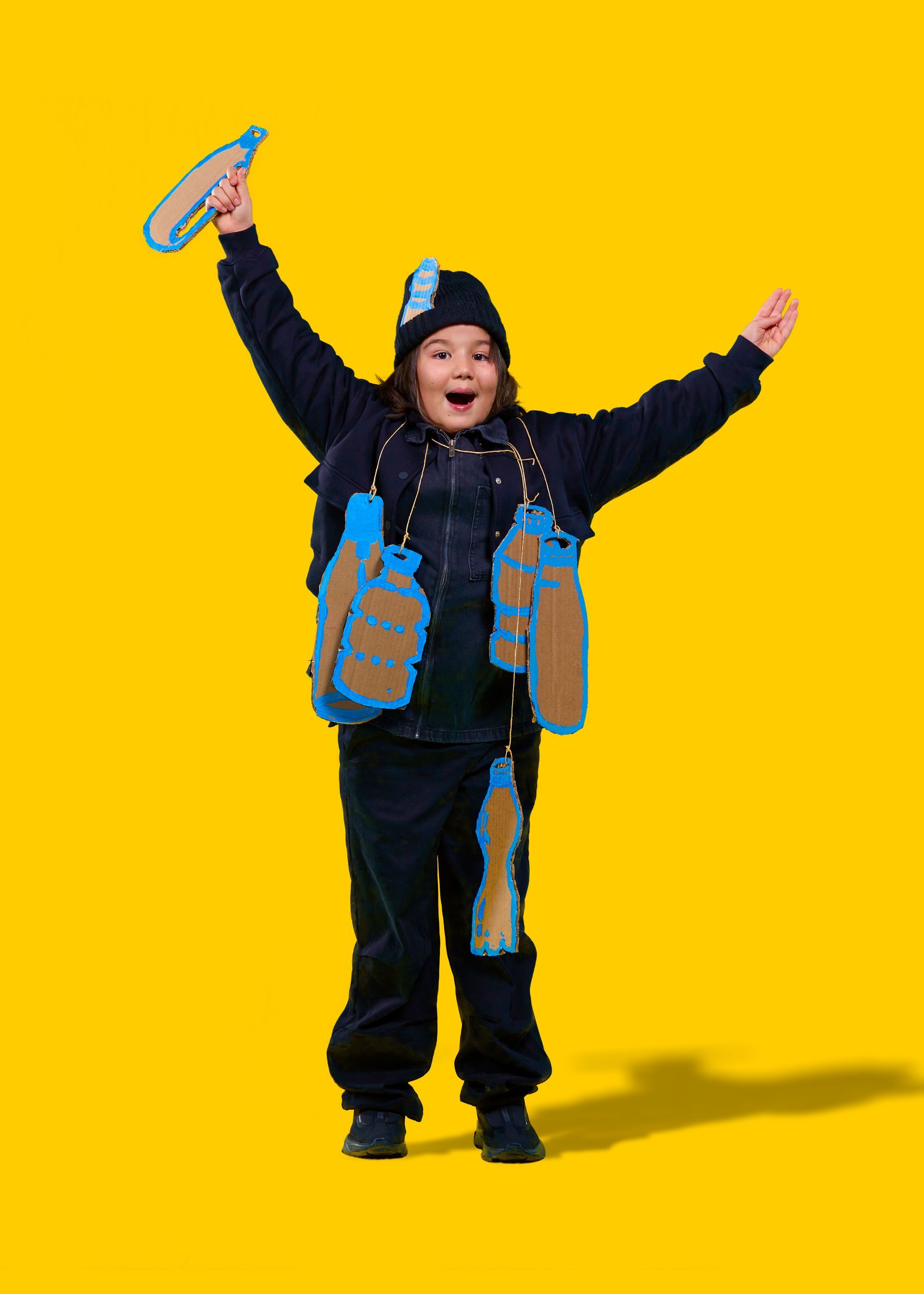 Kind in donkerblauwe outfit, muts en vest versierd met waterflessen, armen omhoog, feestvierend. Gele achtergrond.